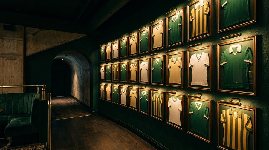 Dinding penuh jersey bola terbingkai di ruang loung stadion yang gelap dengan sorotan cahaya keemasan